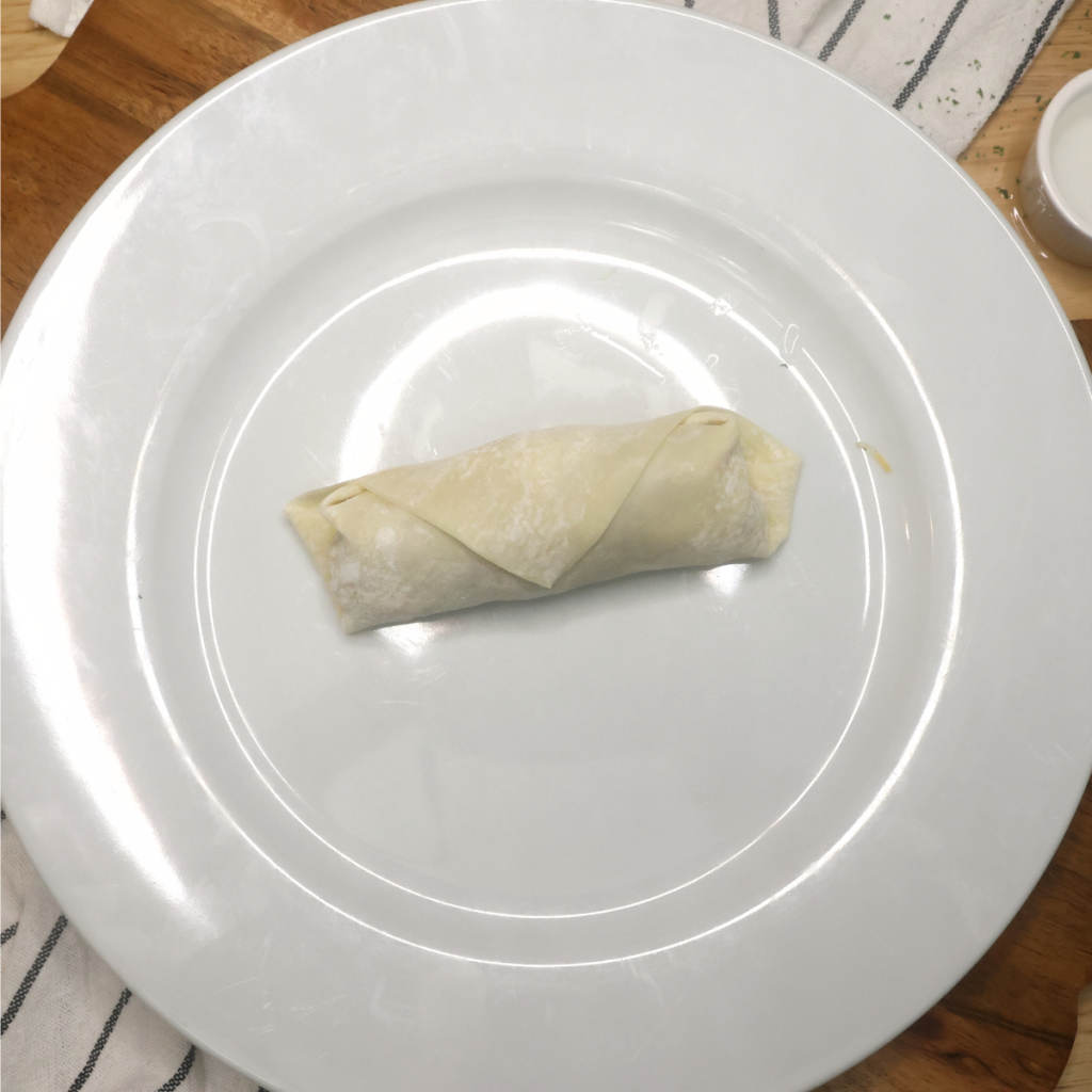 An uncooked cheeseburger egg roll rolled up on a white plate